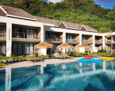 Two-story modern white villa with a tiled roof and glass balcony doors overlooks a turquoise swimming pool with colorful umbrellas; lush green trees create a calm, upscale resort feel.