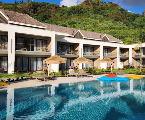Two-story modern white villa with a tiled roof and glass balcony doors overlooks a turquoise swimming pool with colorful umbrellas; lush green trees create a calm, upscale resort feel.