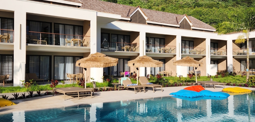 Two-story modern white villa with a tiled roof and glass balcony doors overlooks a turquoise swimming pool with colorful umbrellas; lush green trees create a calm, upscale resort feel.