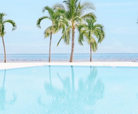 A bright blue infinity pool with reflections of palm trees overlooks a sandy beach and the ocean under a clear sky.