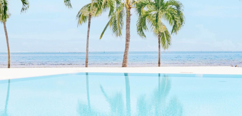 A bright blue infinity pool with reflections of palm trees overlooks a sandy beach and the ocean under a clear sky.