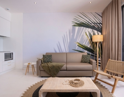 Minimal modern living room with a gray sectional sofa, olive throw, palm fronds, and a woven coffee table set on a rug; muted tones create a calm, cozy mood.