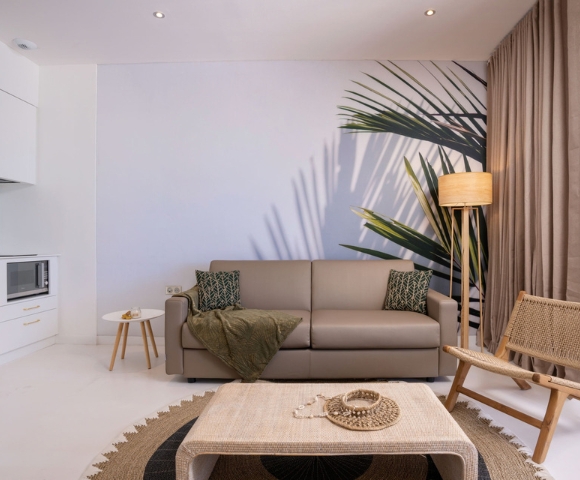 Minimal modern living room with a gray sectional sofa, olive throw, palm fronds, and a woven coffee table set on a rug; muted tones create a calm, cozy mood.