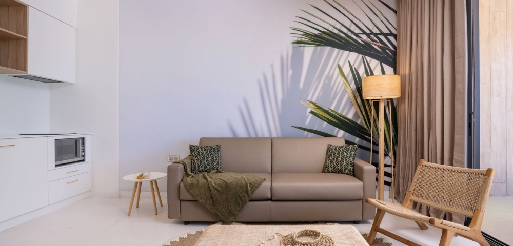 Minimal modern living room with a gray sectional sofa, olive throw, palm fronds, and a woven coffee table set on a rug; muted tones create a calm, cozy mood.