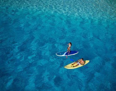 Two people enjoy water sports in crystal clear turquoise water; one stands on a paddleboard, the other sits in a yellow kayak.