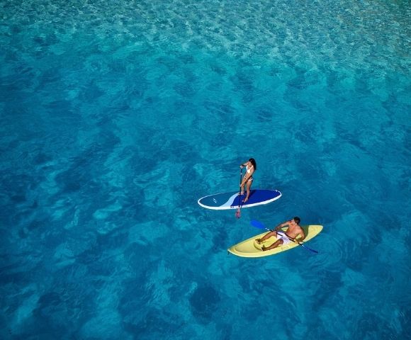 Two people enjoy water sports in crystal clear turquoise water; one stands on a paddleboard, the other sits in a yellow kayak.