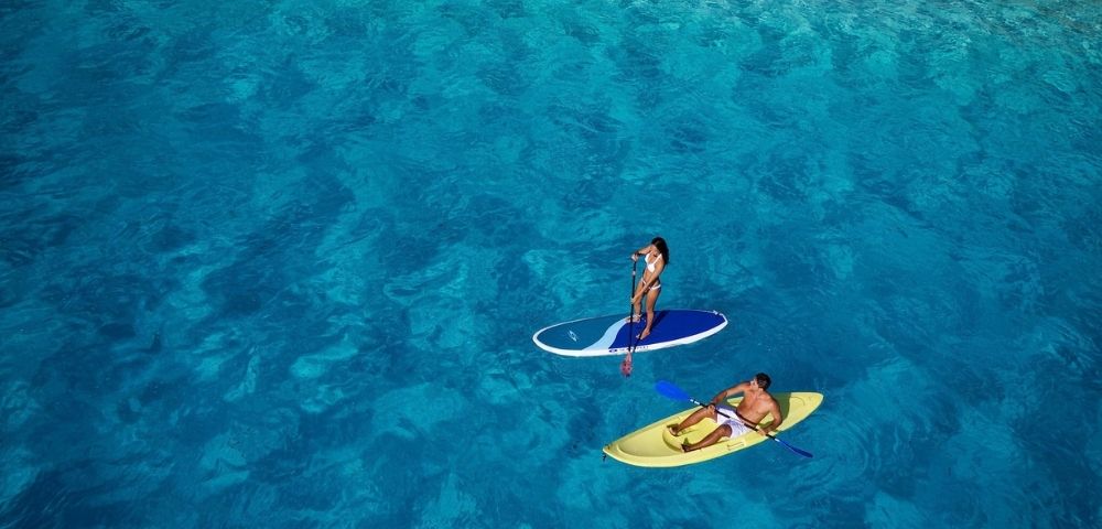Two people enjoy water sports in crystal clear turquoise water; one stands on a paddleboard, the other sits in a yellow kayak.