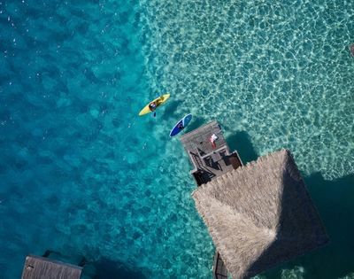 Aerial view of a yellow kayak and a blue paddleboard floating on clear turquoise water near a wooden dock and a thatched-roof bungalow over the water.