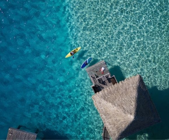 Aerial view of a yellow kayak and a blue paddleboard floating on clear turquoise water near a wooden dock and a thatched-roof bungalow over the water.