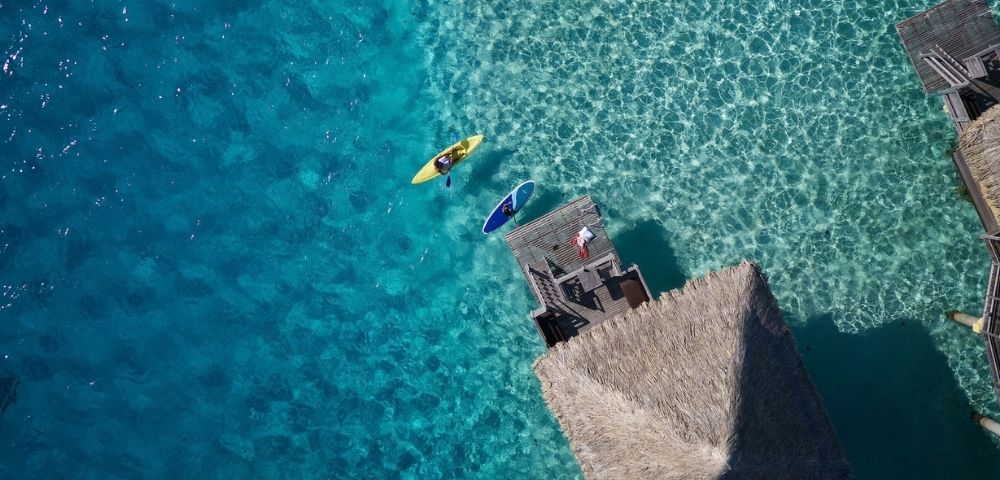 Aerial view of a yellow kayak and a blue paddleboard floating on clear turquoise water near a wooden dock and a thatched-roof bungalow over the water.