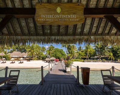 Wooden pier with a thatched roof overhang leading to a sandy beach lined with palm trees and resort buildings.