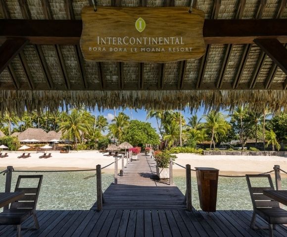 Wooden pier with a thatched roof overhang leading to a sandy beach lined with palm trees and resort buildings.
