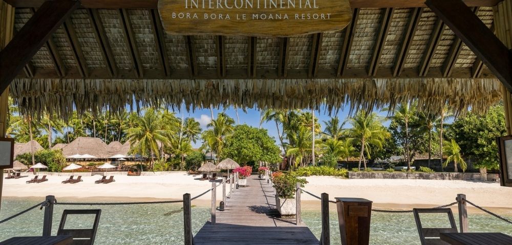 Wooden pier with a thatched roof overhang leading to a sandy beach lined with palm trees and resort buildings.