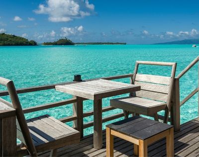 Wooden chairs and tables on a deck overlooking a vibrant turquoise ocean with distant islands under a bright blue sky with white clouds.