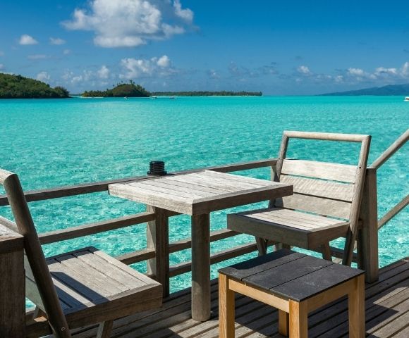 Wooden chairs and tables on a deck overlooking a vibrant turquoise ocean with distant islands under a bright blue sky with white clouds.