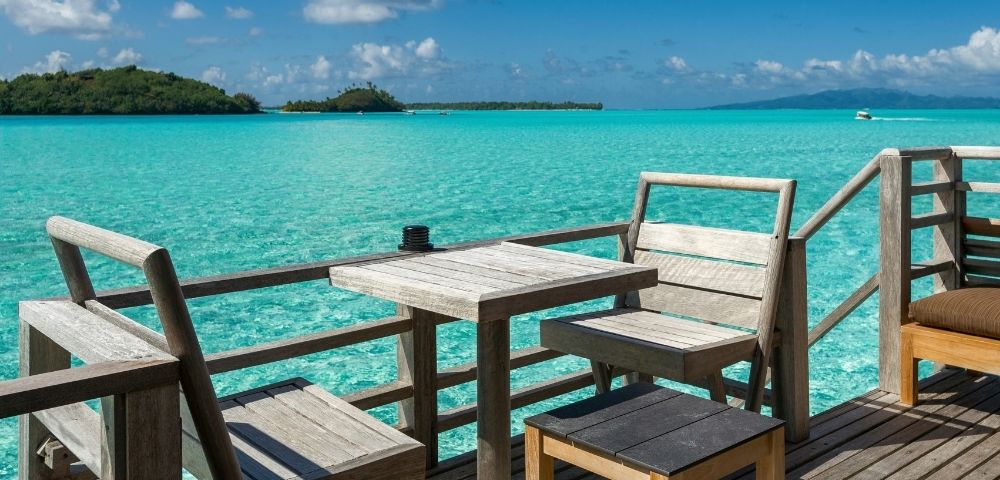 Wooden chairs and tables on a deck overlooking a vibrant turquoise ocean with distant islands under a bright blue sky with white clouds.