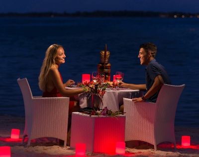 A couple enjoys a romantic dinner on the beach at night, with a tiki statue centerpiece and red lighting.