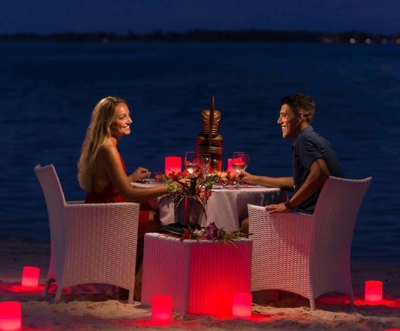 A couple enjoys a romantic dinner on the beach at night, with a tiki statue centerpiece and red lighting.