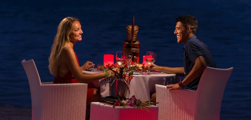 A couple enjoys a romantic dinner on the beach at night, with a tiki statue centerpiece and red lighting.