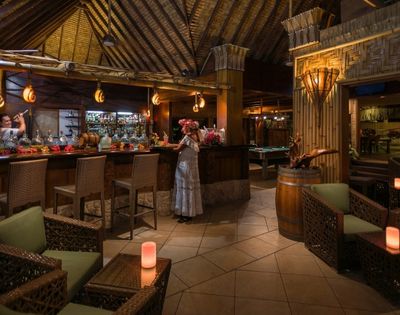 A woman stands at a rustic tropical bar beneath a thatched timber roof, surrounded by wicker seating and lanterns in warm, inviting light.