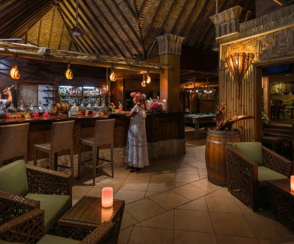 A woman stands at a rustic tropical bar beneath a thatched timber roof, surrounded by wicker seating and lanterns in warm, inviting light.
