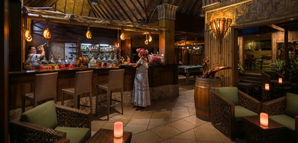 A woman stands at a rustic tropical bar beneath a thatched timber roof, surrounded by wicker seating and lanterns in warm, inviting light.