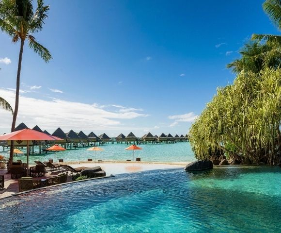 A tranquil tropical resort with a turquoise infinity pool, palm trees, red umbrellas, and overwater bungalows beneath a bright blue sky.