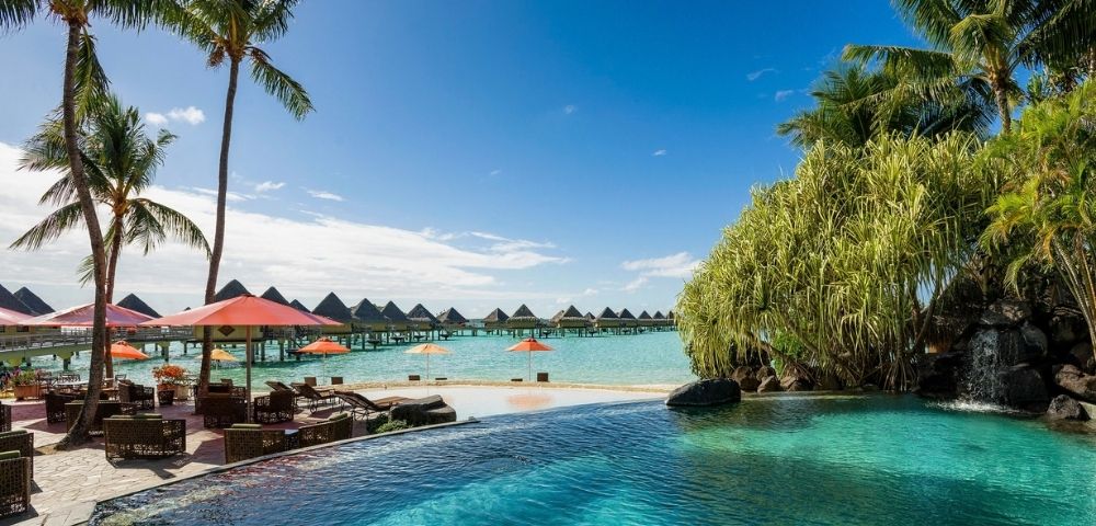 A tranquil tropical resort with a turquoise infinity pool, palm trees, red umbrellas, and overwater bungalows beneath a bright blue sky.