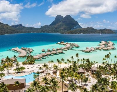Aerial view of a tropical island resort with overwater bungalows, turquoise lagoon, palm-lined beach, and dramatic green mountains under a bright blue sky.