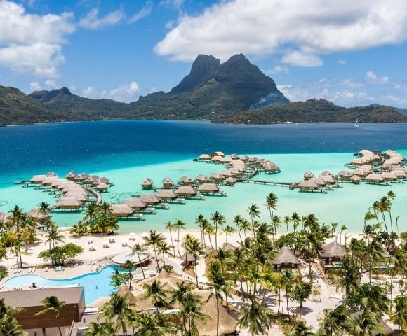 Aerial view of a tropical island resort with overwater bungalows, turquoise lagoon, palm-lined beach, and dramatic green mountains under a bright blue sky.