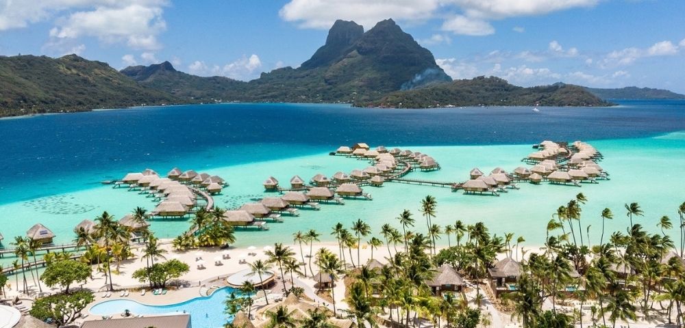 Aerial view of a tropical island resort with overwater bungalows, turquoise lagoon, palm-lined beach, and dramatic green mountains under a bright blue sky.