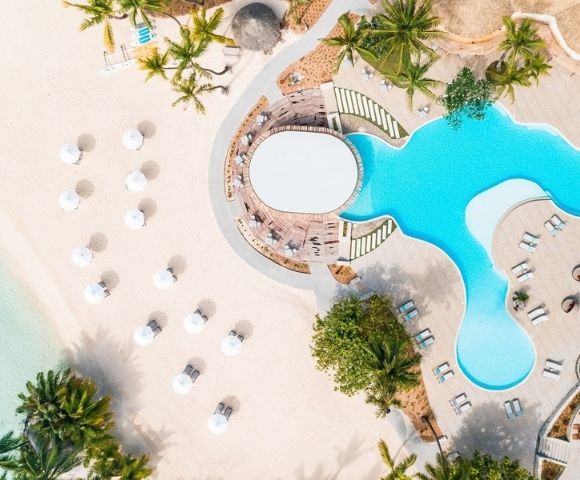 Aerial view of a turquoise resort pool beside a sandy beach, with white umbrellas, sun loungers and palm trees creating a serene tropical escape.