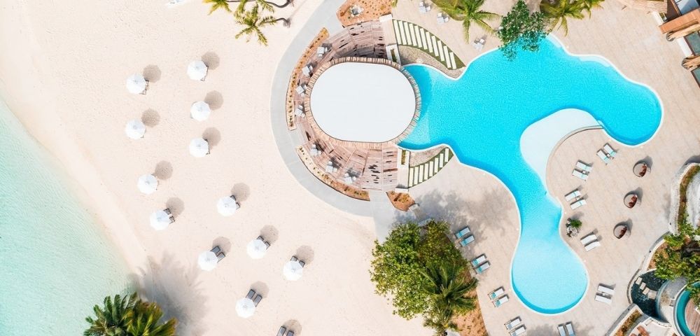 Aerial view of a turquoise resort pool beside a sandy beach, with white umbrellas, sun loungers and palm trees creating a serene tropical escape.