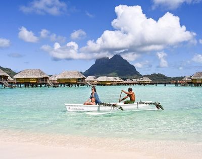 Two people paddle an outrigger canoe across turquoise lagoon water, with overwater bungalows and a lush mountain beneath bright clouds, serene tropical scene.