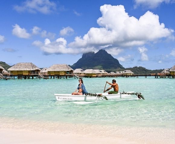 Two people paddle an outrigger canoe across turquoise lagoon water, with overwater bungalows and a lush mountain beneath bright clouds, serene tropical scene.