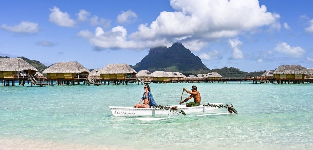Two people paddle an outrigger canoe across turquoise lagoon water, with overwater bungalows and a lush mountain beneath bright clouds, serene tropical scene.