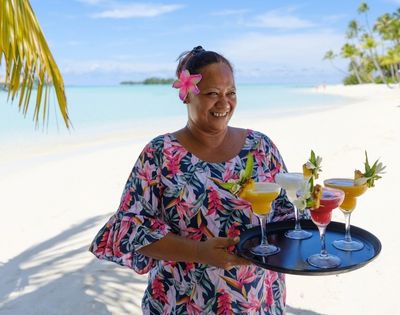 A smiling woman with a pink flower in her hair holds colorful tropical cocktails on a sunny white-sand beach, conveying relaxed island warmth.