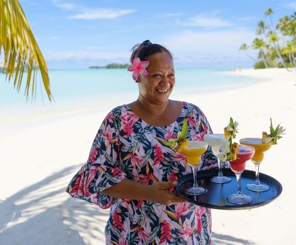 A smiling woman with a pink flower in her hair holds colorful tropical cocktails on a sunny white-sand beach, conveying relaxed island warmth.