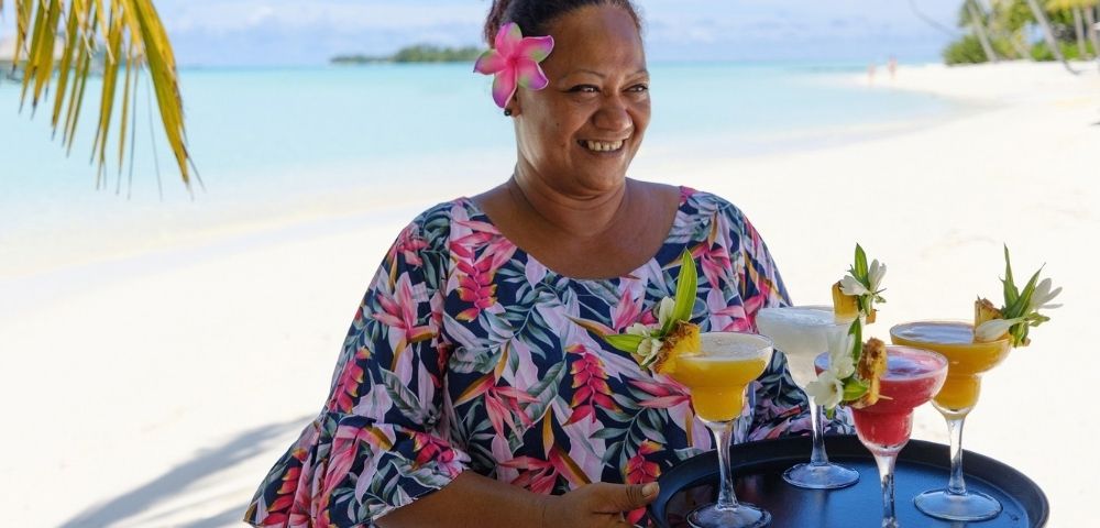A smiling woman with a pink flower in her hair holds colorful tropical cocktails on a sunny white-sand beach, conveying relaxed island warmth.