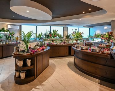 A modern buffet restaurant with curved wooden food counters, colorful dishes, tropical plants, and large windows, creating a fresh, welcoming atmosphere.