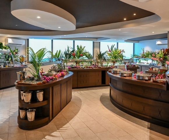 A modern buffet restaurant with curved wooden food counters, colorful dishes, tropical plants, and large windows, creating a fresh, welcoming atmosphere.