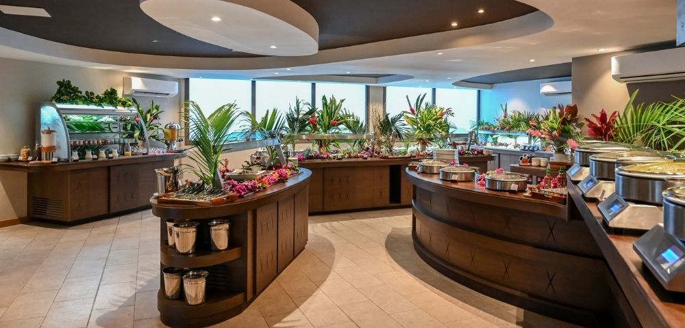 A modern buffet restaurant with curved wooden food counters, colorful dishes, tropical plants, and large windows, creating a fresh, welcoming atmosphere.