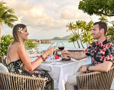 A couple in tropical resort attire toast with wine at a poolside table, surrounded by palm trees and ocean views in a relaxed, romantic setting.