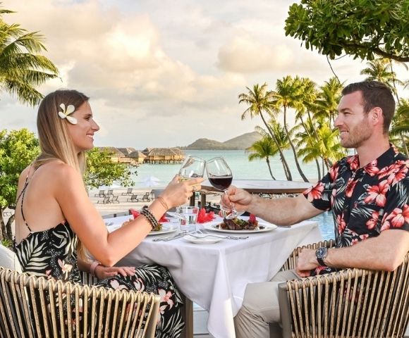 A couple in tropical resort attire toast with wine at a poolside table, surrounded by palm trees and ocean views in a relaxed, romantic setting.