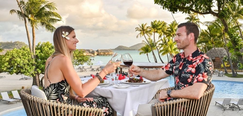 A couple in tropical resort attire toast with wine at a poolside table, surrounded by palm trees and ocean views in a relaxed, romantic setting.