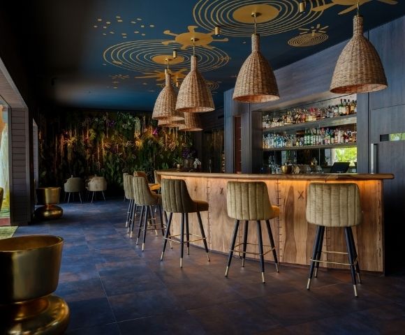 Warm, upscale bar interior with a glowing wood counter, upholstered stools, woven pendant lights, and a stocked back bar, creating a relaxed, intimate mood.