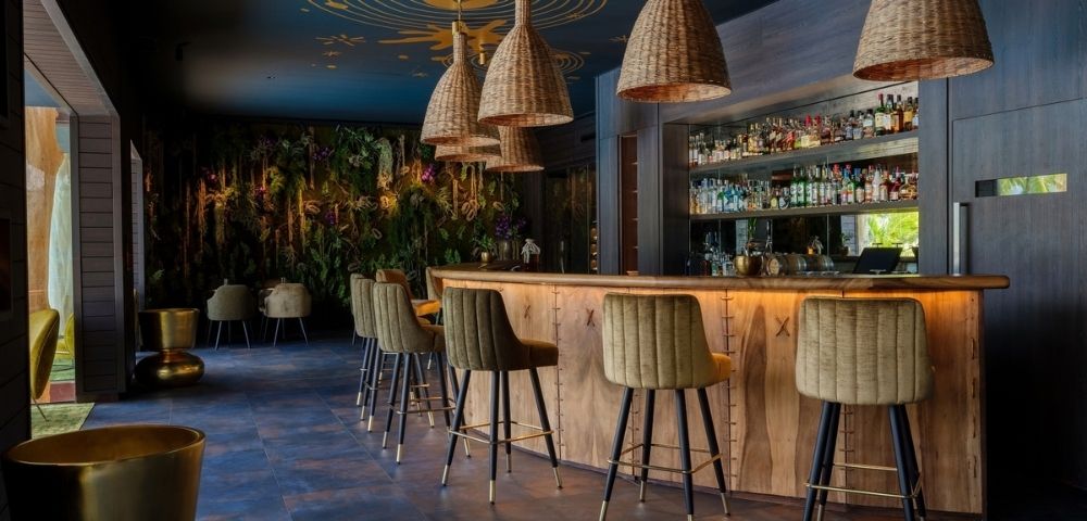Warm, upscale bar interior with a glowing wood counter, upholstered stools, woven pendant lights, and a stocked back bar, creating a relaxed, intimate mood.