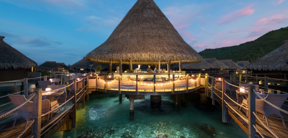 A thatched-roof overwater bar and restaurant at twilight, with illuminated walkways and tables, set in clear turquoise water where fish swim.
