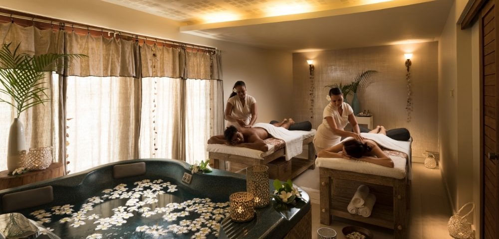 A serene spa treatment room featuring two massage tables with therapists giving massages to clients, a large, bubbling jacuzzi filled with floating white flowers, and ambient lighting.