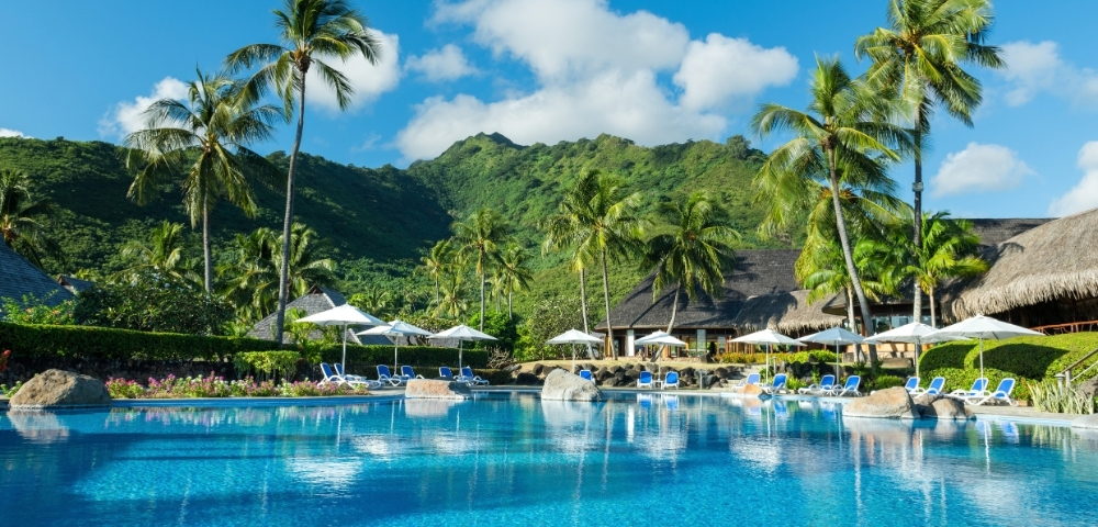 A bright blue resort swimming pool with lounge chairs and umbrellas, surrounded by lush green palm trees and a tropical mountain backdrop under a clear blue sky.
